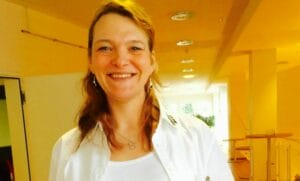 A smiling woman in a white shirt standing in a hallway.