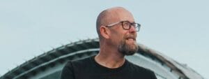 A bald man with glasses is standing in front of a building.