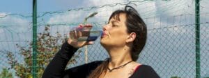 A woman drinking water from a bottle in front of a fence.