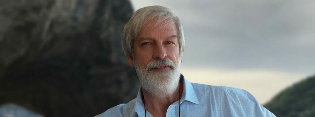 A man with a beard and white hair standing in front of a mountain.