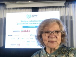 An older woman takes a selfie in front of a presentation screen at a conference. The screen displays logos and text in Slovak.