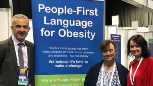 Three people standing in front of a sign that says people first language for obesity.