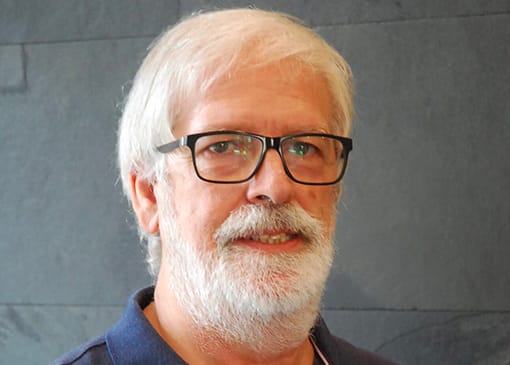 A man with glasses and a beard standing in front of a wall.