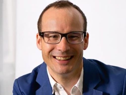 A smiling man wearing glasses and a blue jacket.