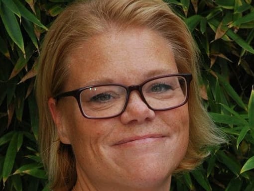 A woman in glasses is smiling in front of a bush.