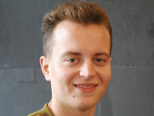 A young man smiling in front of a gray wall.