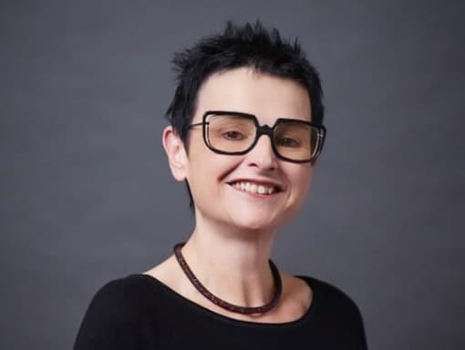 A woman in glasses smiling at a grey background.