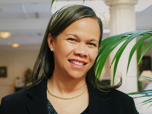 A woman in a black suit smiles in front of a plant.