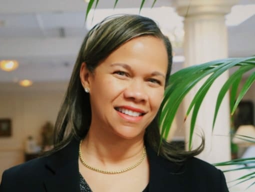A woman in a black suit smiles in front of a plant.