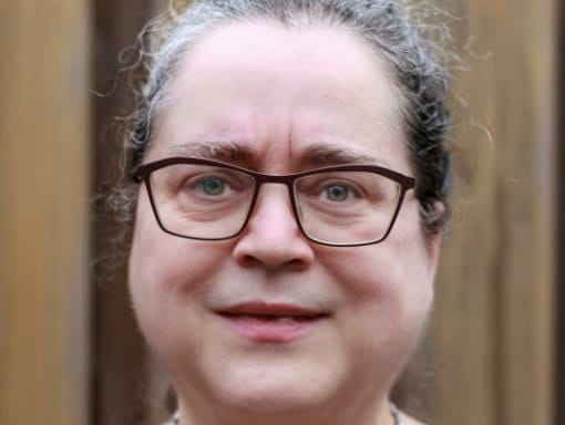 Close-up portrait of a middle-aged woman with curly gray hair, wearing glasses, and slightly smiling against a wooden background.