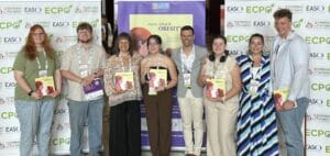 Eight people stand in front of banners and a poster at an event. They are holding up booklets, smiling, and wearing event badges.