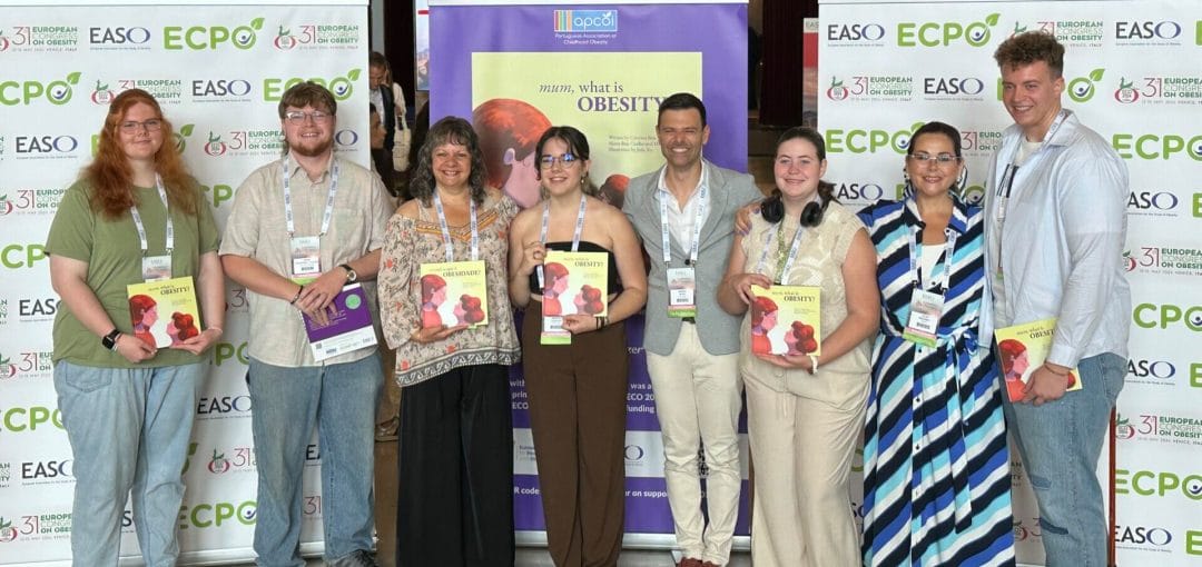 Eight people stand in front of banners and a poster at an event. They are holding up booklets, smiling, and wearing event badges.
