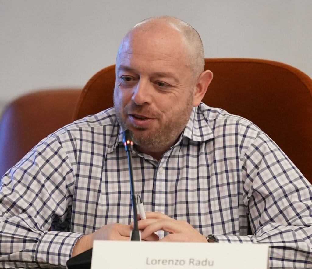A man in a checkered shirt speaks into a microphone at a conference table. A nameplate in front of him reads "Lorenzo Radu.