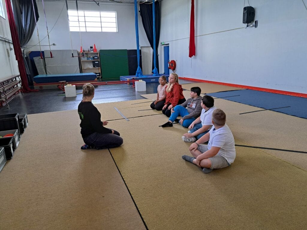 A group of children sits in a circle on a gym floor, listening to an instructor wearing a black shirt. Gymnastics equipment is visible in the background.