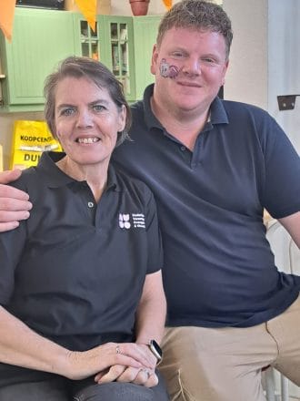 Two people wearing black shirts sit together smiling. The man has a painted design on his cheek. Orange decorations are visible in the background.