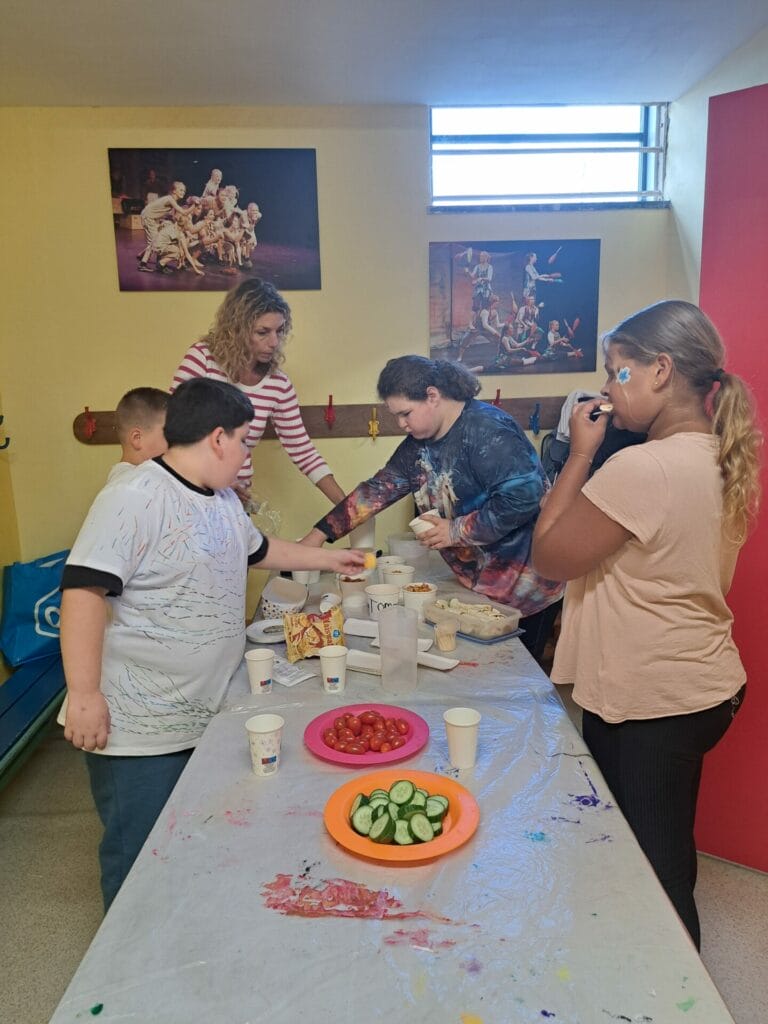A group of children and an adult are preparing food at a table with paints and craft materials. Plates of sliced cucumbers and tomatoes are visible. Posters hang on the wall above them.