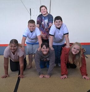 Six people forming a human pyramid indoors, three on the bottom, two in the middle, and one on top, all smiling.
