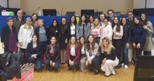 Group photo of 23 people in a room, standing and smiling. Some are dressed in formal attire, while others are in casual clothing.