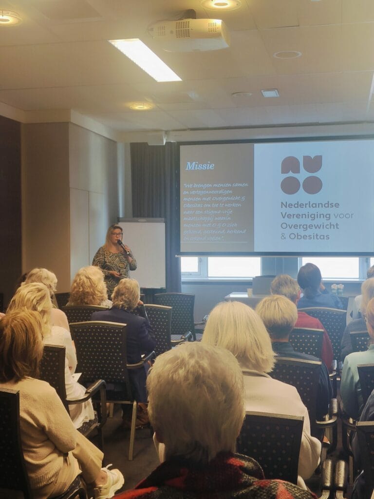 A woman presents to an audience in a conference room; a slide shows the logo and mission of the Nederlandse Vereniging voor Overgewicht & Obesitas.