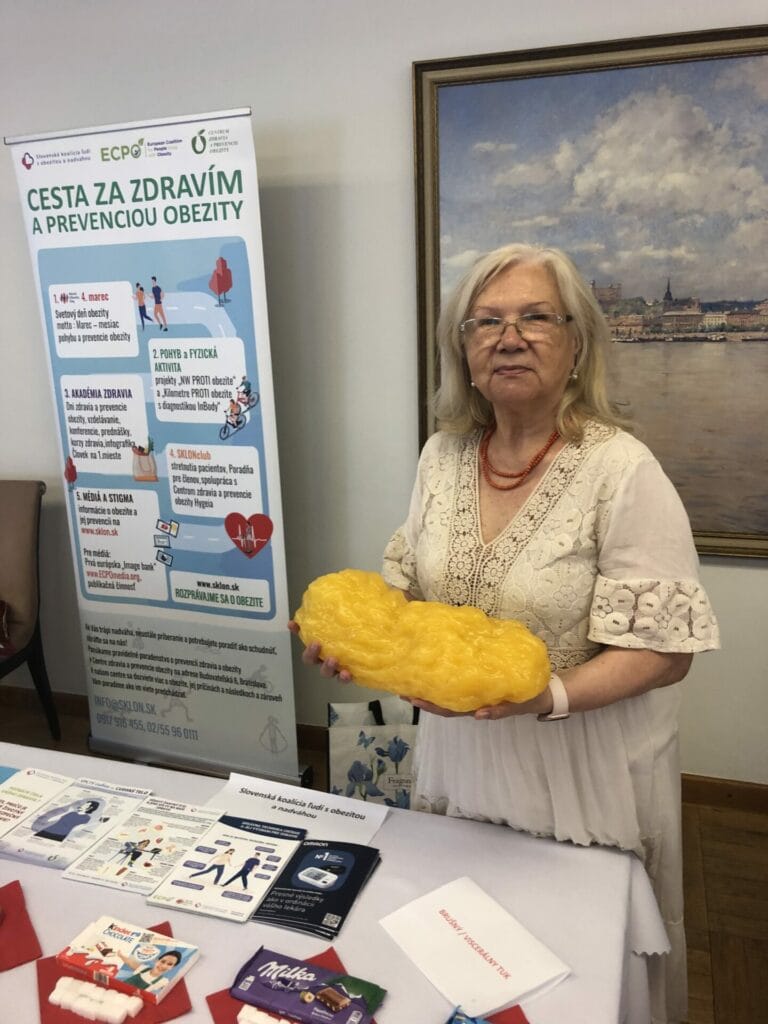 An elderly woman holds a large model of body fat. Behind her is a poster about obesity prevention in Slovak and a table with informational pamphlets.