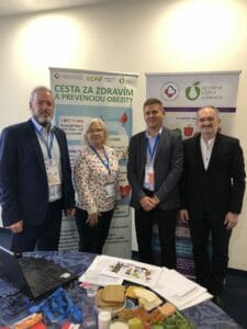 Four people stand in front of banners promoting health and obesity prevention. A table with health-related materials and food items is in the foreground.