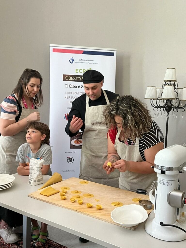 People in aprons making pasta at a table with a pasta sheet and plates. A boy is watching, and a poster is in the background.