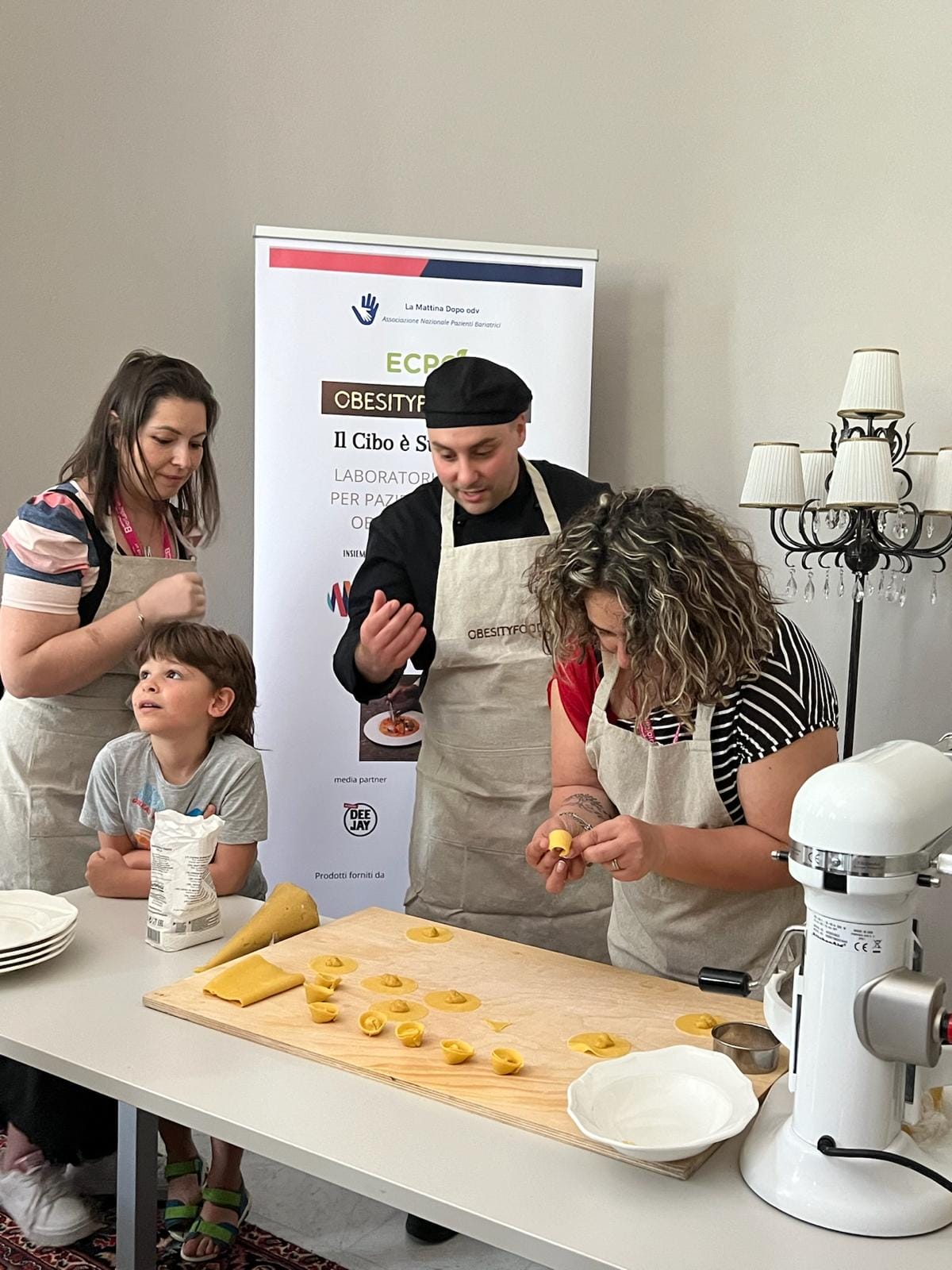 People in aprons making pasta at a table with a pasta sheet and plates. A boy is watching, and a poster is in the background.