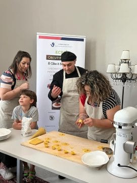 People in aprons making pasta at a table with a pasta sheet and plates. A boy is watching, and a poster is in the background.