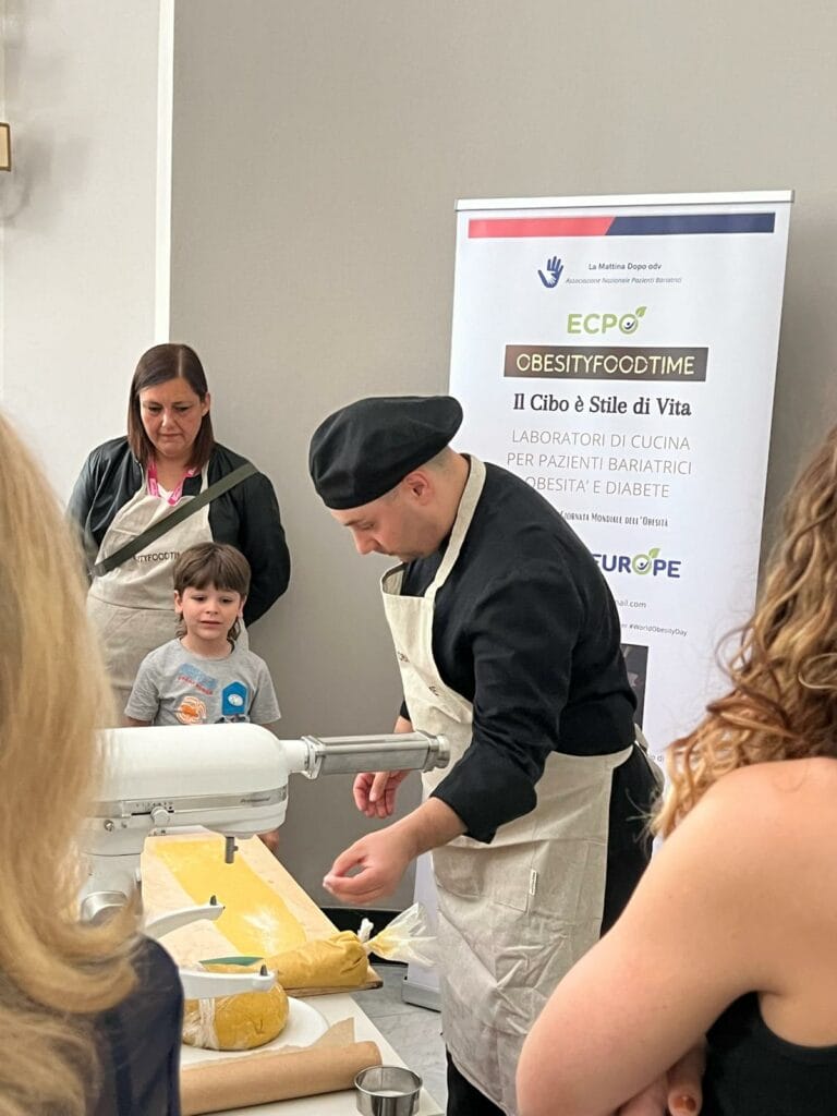 A chef in black attire prepares dough on a table while a woman and child observe. A banner in the background mentions cooking workshops for bariatric patients.