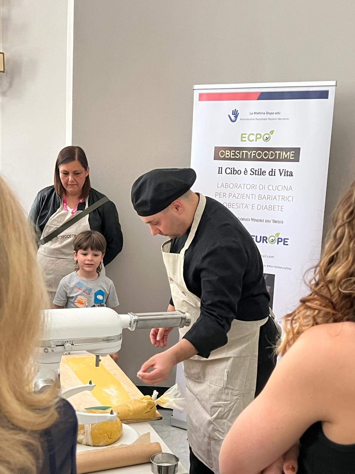 A chef in black attire prepares dough on a table while a woman and child observe. A banner in the background mentions cooking workshops for bariatric patients.