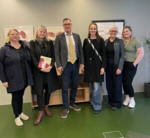 A group of six people standing together indoors, dressed in professional and casual attire, with framed art and a wooden cabinet in the background.