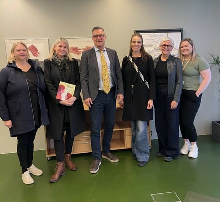 A group of six people standing together indoors, dressed in professional and casual attire, with framed art and a wooden cabinet in the background.