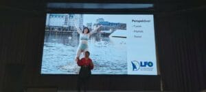 A person stands on stage in front of a large screen displaying a woman in a swimsuit in a city waterfront setting. The screen includes Norwegian text and a logo at the bottom.