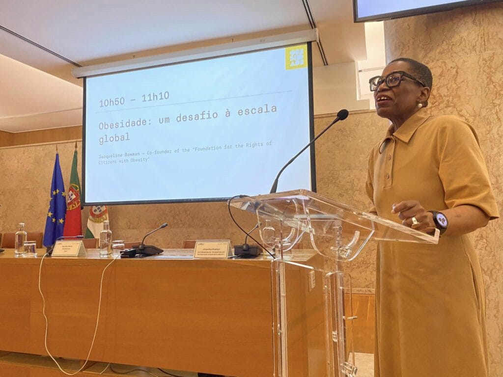 A person standing at a podium giving a presentation titled "Obesidade: um desafio à escala global" with flags and a presentation screen in the background.
