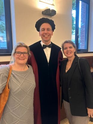 Three people posing indoors; one is wearing academic attire with a hat and robe, while the other two stand beside in casual clothes, smiling.