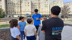 A group of children and a man in a blue shirt are gathered outdoors in an urban area. Another person in a black shirt with "Gasol Foundation" on the back stands nearby.