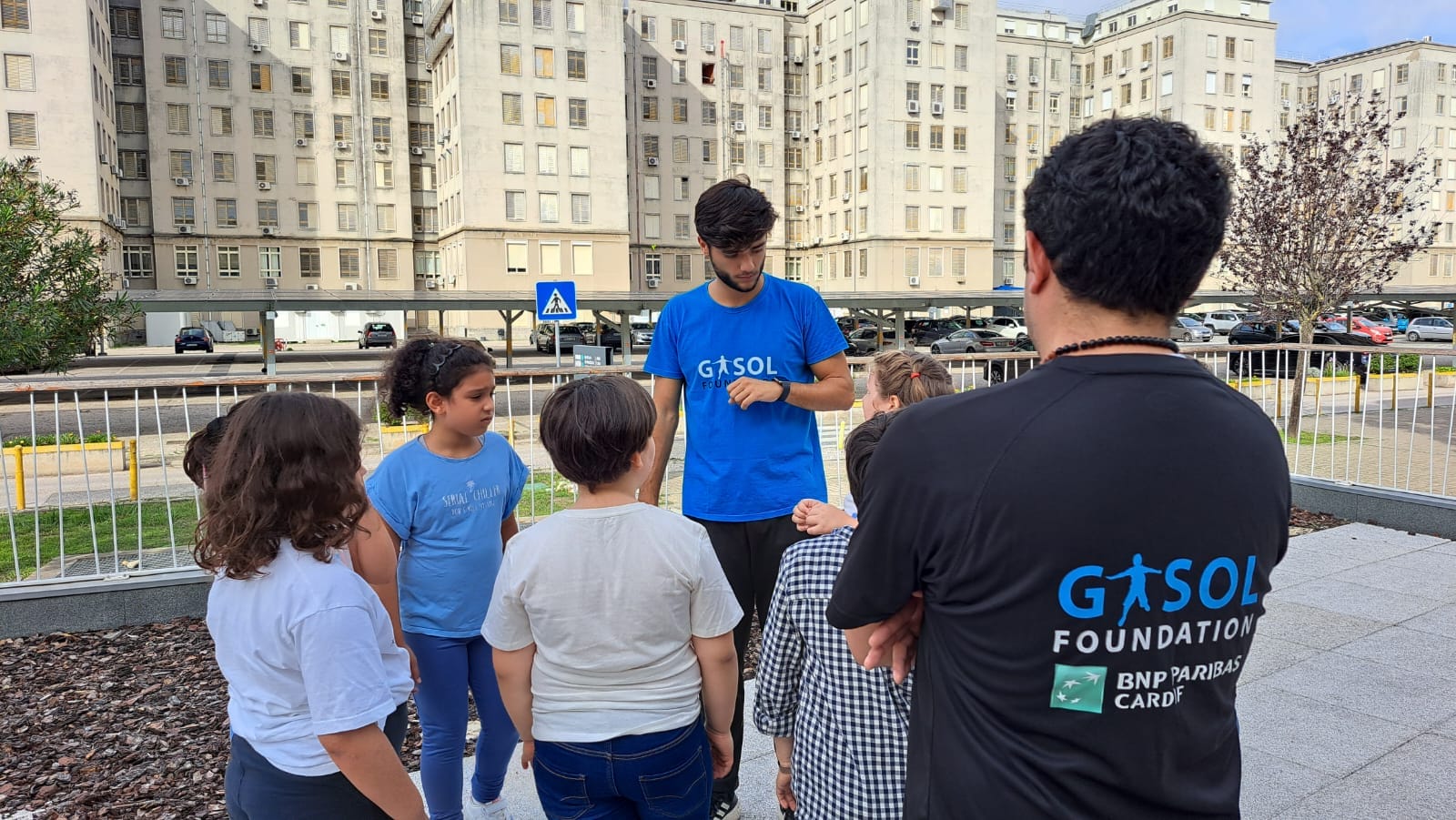 A group of children and a man in a blue shirt are gathered outdoors in an urban area. Another person in a black shirt with "Gasol Foundation" on the back stands nearby.