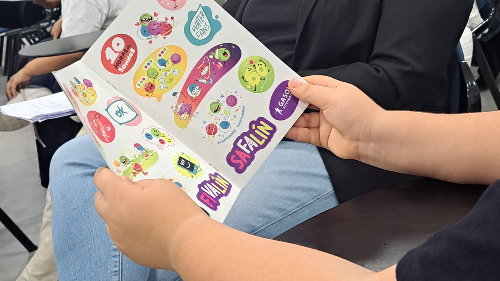 A person holds a sheet of colorful stickers with various designs while seated in a room with others.