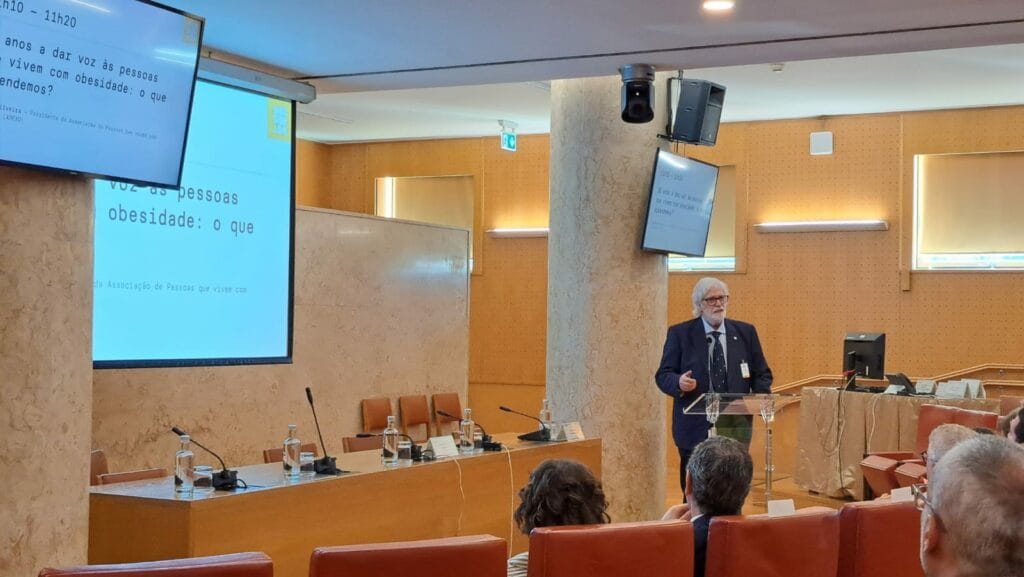 A speaker stands at a podium in a conference room, addressing an audience. A presentation is displayed on screens behind and above.