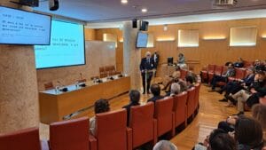 A man speaks at a podium in a conference room. The audience is seated, and a presentation is displayed on a screen behind him.
