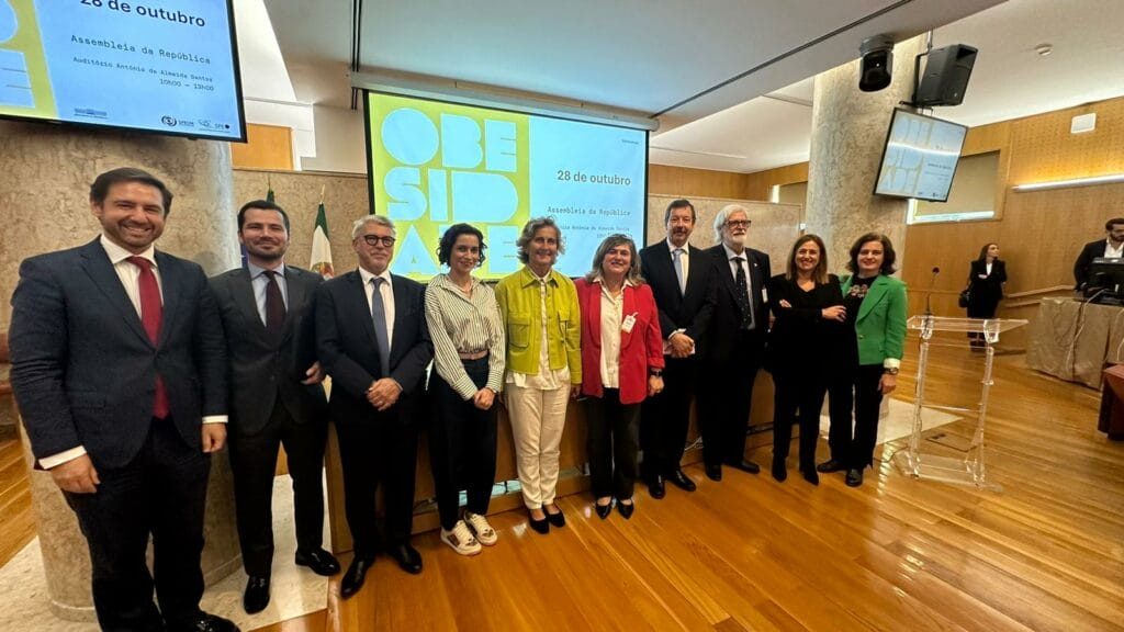 A group of people stands in a room with a wood floor and screen displaying the date "28 de outubro.