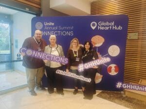 Four people stand in front of a conference backdrop holding signs with hashtags and slogans related to the event.