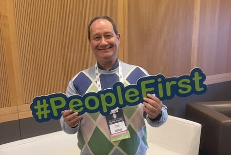 A person smiling, holding a "#PeopleFirst" sign indoors, wearing a patterned sweater and event badge.