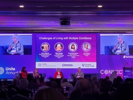 Panel discussion at a conference on "Challenges of Living with Multiple Conditions," featuring four speakers on stage with their images displayed on a large screen. Audience seated in foreground.