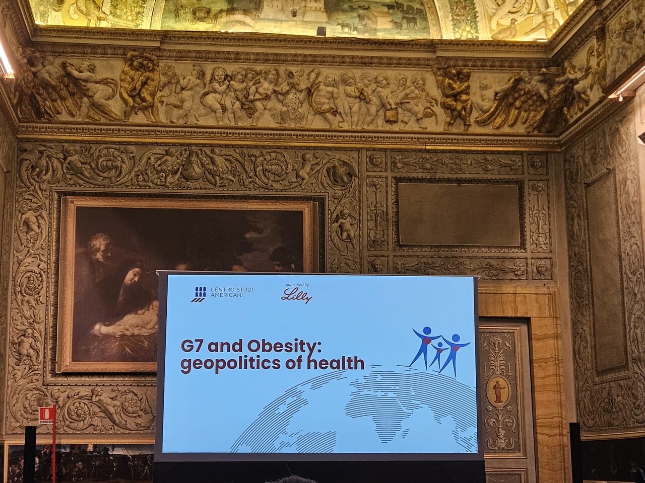 A conference room with ornate walls and ceiling, featuring a large screen displaying "G7 and Obesity: geopolitics of health" and logos of Centro Studi Americani and Lilly.