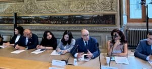A group of people in formal attire sit at a long table in a decorated room with large paintings on the walls. One man is speaking and gesturing with his hands.
