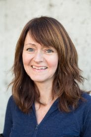 A person with shoulder-length brown hair and a blue top smiles while standing against a light-colored background.