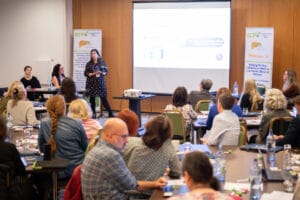 A woman presents in front of a seated audience at a conference, standing beside a screen displaying graphs and text.