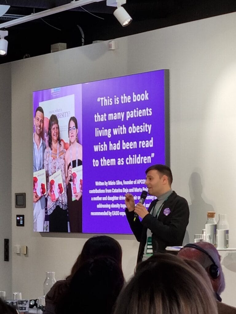 A person presenting in front of an audience with a slide showing three people holding books and a quote about a book on obesity.