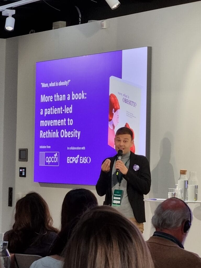 Person giving a presentation in front of a screen displaying a graphic about a patient-led movement to rethink obesity. Audience members are seated.